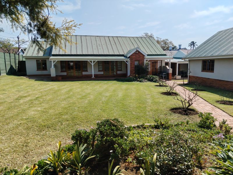 A well-maintained rear exterior view of the house featuring a spacious lawn, brick paved pathways, and a covered patio area with timber framed doors.