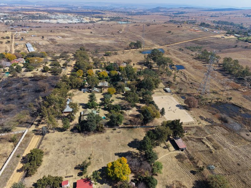 Aerial view of a large rural property featuring mature trees, open land, a sand arena, and multiple outbuildings set within a vast landscape.