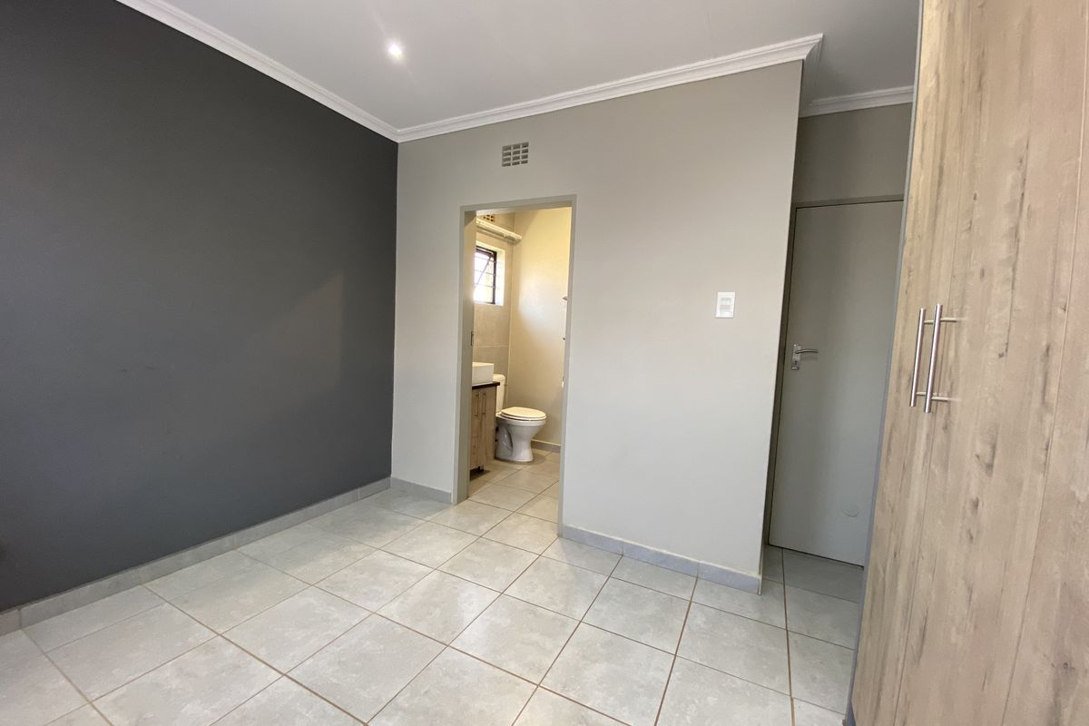 A clean, empty bedroom featuring tiled flooring, built-in cupboards, and an en-suite bathroom access point with a modern neutral and dark grey color scheme.