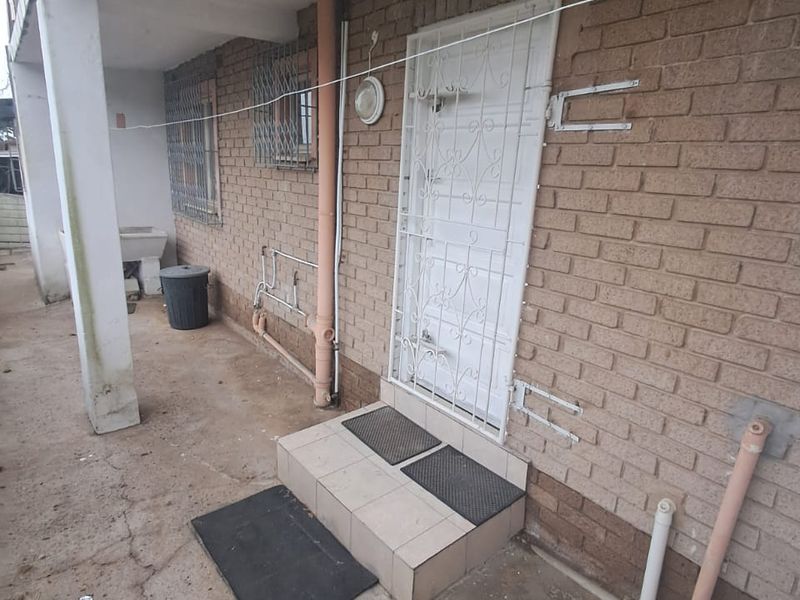 Rear exterior area of the cottage featuring a brick facade, security gate, concrete patio, and external utility plumbing.