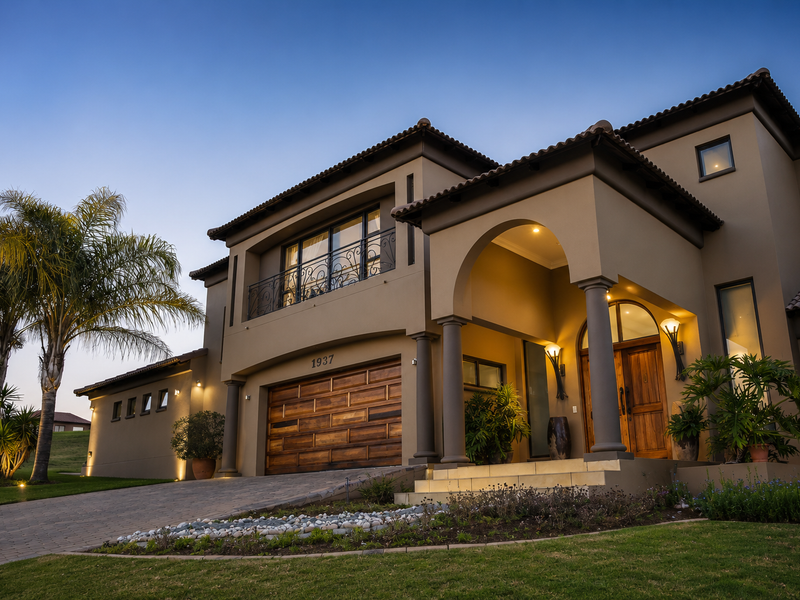 Stunning double-story residence featuring an arched portico, stone columns, a wooden garage door, and a beautifully landscaped front garden at dusk.
