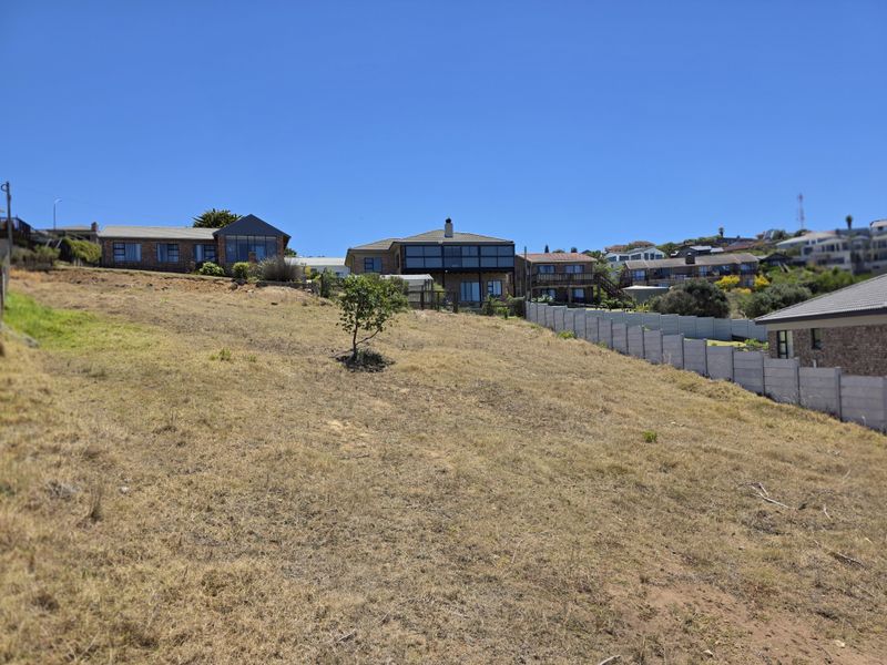 A view of a vacant residential plot with a gentle slope, featuring surrounding neighborhood homes and a precast concrete boundary wall.