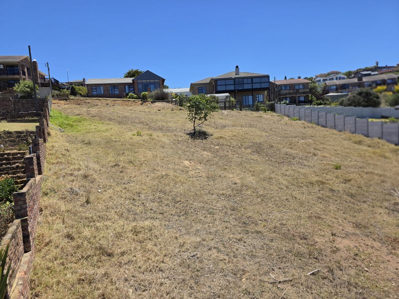 A vacant residential plot featuring a sloped landscape, bordered by existing brick retaining walls and concrete fencing in a developed neighborhood.