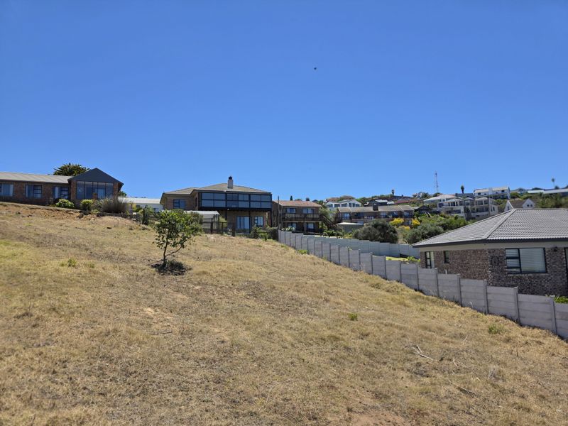 A sloped vacant residential plot featuring dry grass, bordered by a precast concrete boundary wall, with surrounding residential properties visible in the background.
