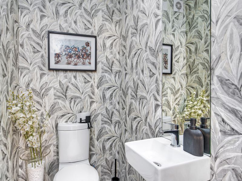 A modern powder room featuring a wall-mounted basin, chrome fixtures, a large mirror, and striking leaf-patterned wallpaper.