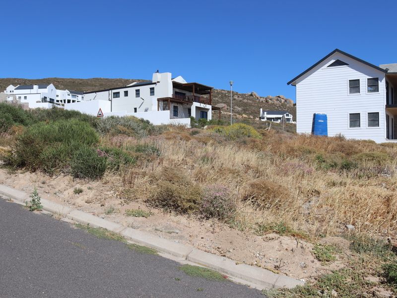 A view of a vacant residential plot featuring sloped terrain, adjacent to a paved road with established neighboring properties in a coastal estate setting.