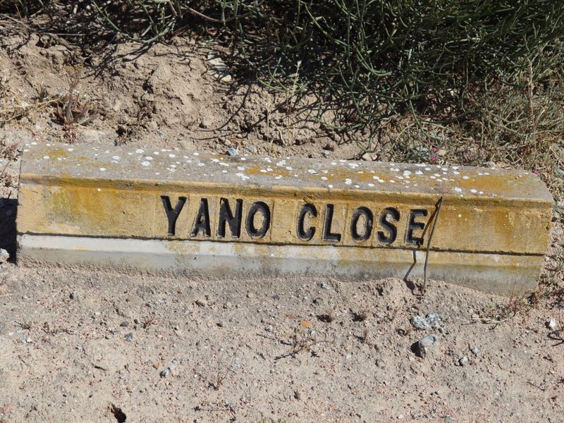 A close-up view of a weathered concrete street name marker for Yano Close located on a sandy verge.
