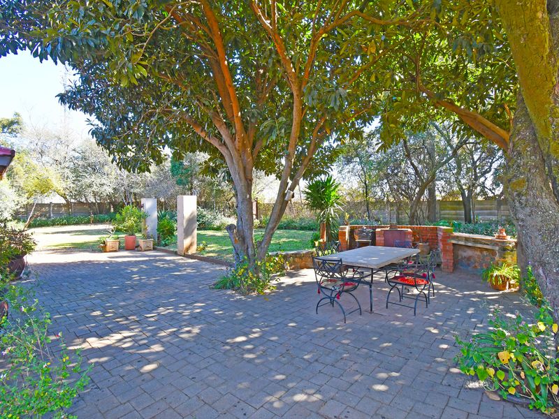A shaded outdoor patio area featuring brick paving, a built-in brick braai, and mature trees, providing a pleasant space for outdoor dining and relaxation.