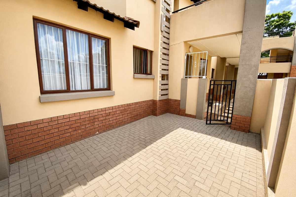 A private paved rear courtyard featuring brick detailing, a security gate, and access to an upper-level balcony.