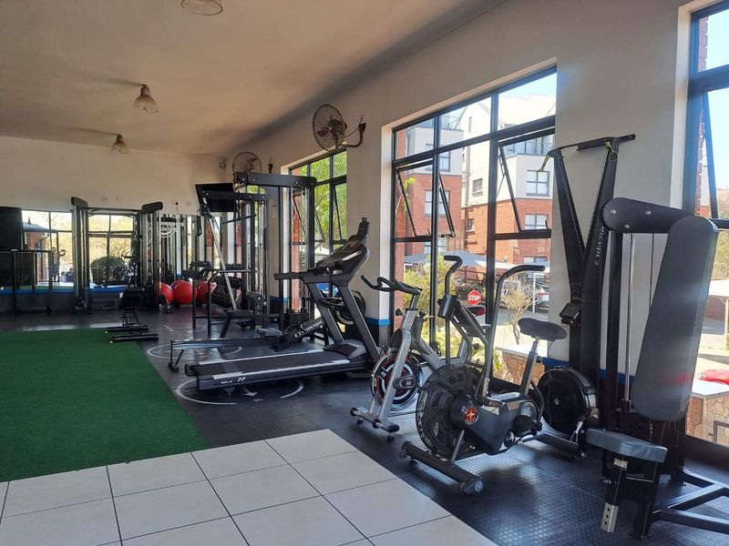 A well-lit, spacious gym facility featuring large windows, a mix of tiled and rubber flooring, and dedicated workout zones.