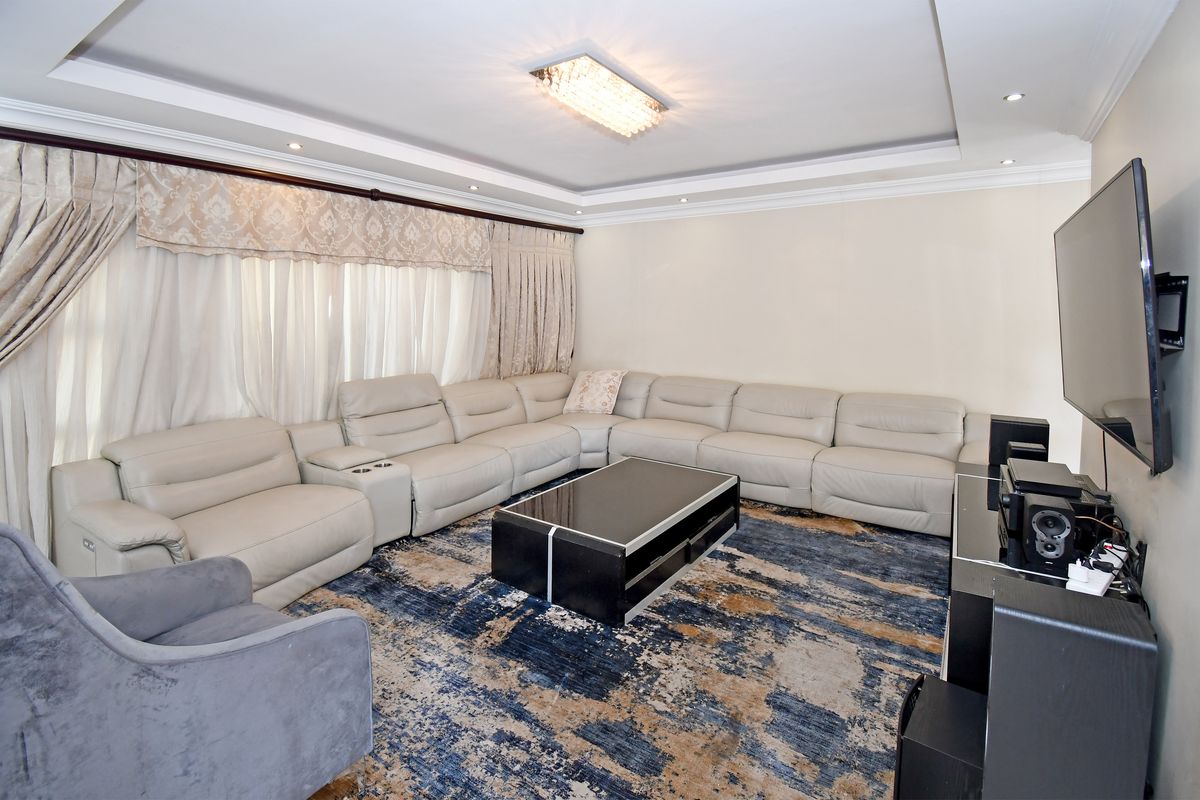 A spacious living room featuring a recessed ceiling with cove lighting, a central crystal chandelier, and elegant curtain pelmets.