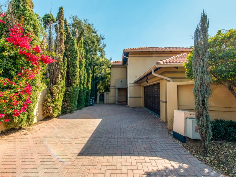 Front exterior view of the property featuring a paved driveway, garage access, and established garden landscaping with mature trees and flowering shrubs.