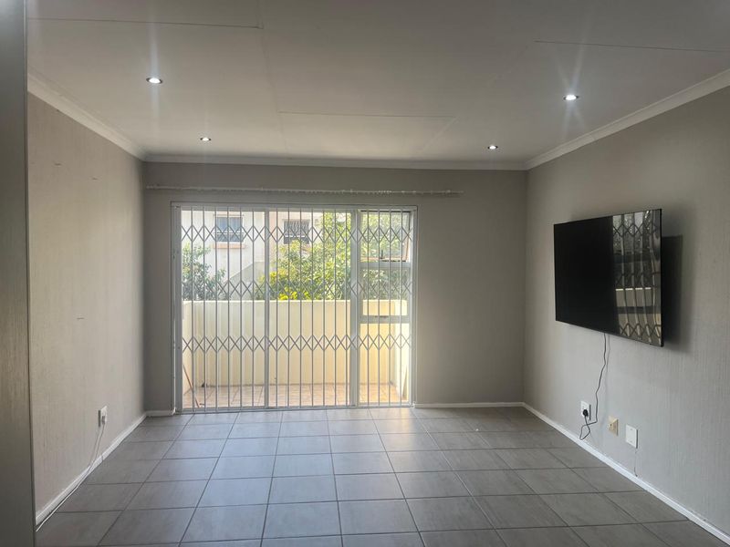 A bright, open-plan living area featuring neutral tiled flooring, recessed downlighting, and a sliding glass door with a security gate leading to a balcony.