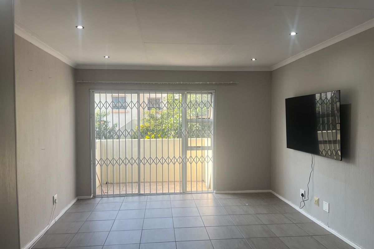A bright, empty living room featuring neutral tiled flooring, recessed downlights, and a sliding glass door with a security gate leading to a balcony.