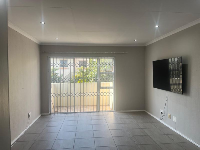 A bright, empty living room featuring neutral tiled flooring, recessed downlights, and a sliding glass door with a security gate leading to a balcony.