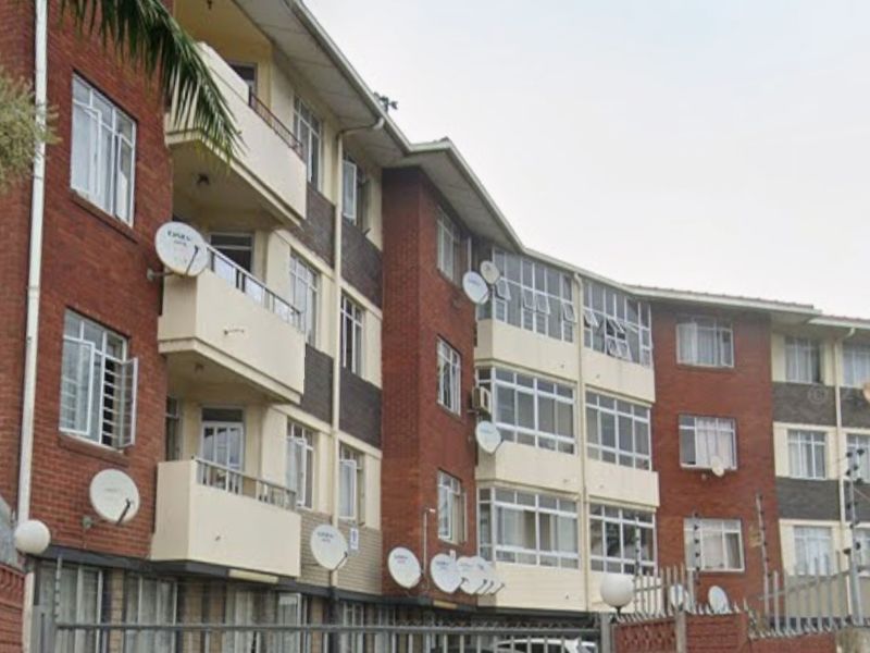 Front elevation of a multi-story brick apartment building featuring balconies, security fencing, and multiple satellite dish installations.