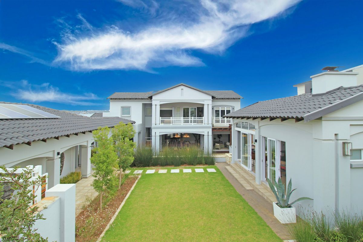 A well-maintained rear exterior view of a modern house featuring a landscaped garden, covered patio, balcony, and solar panels on the tiled roof.