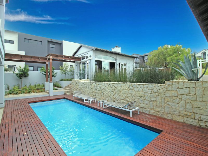A well-maintained outdoor pool area featuring timber decking, a stone feature wall, and a timber pergola, set within a landscaped garden.