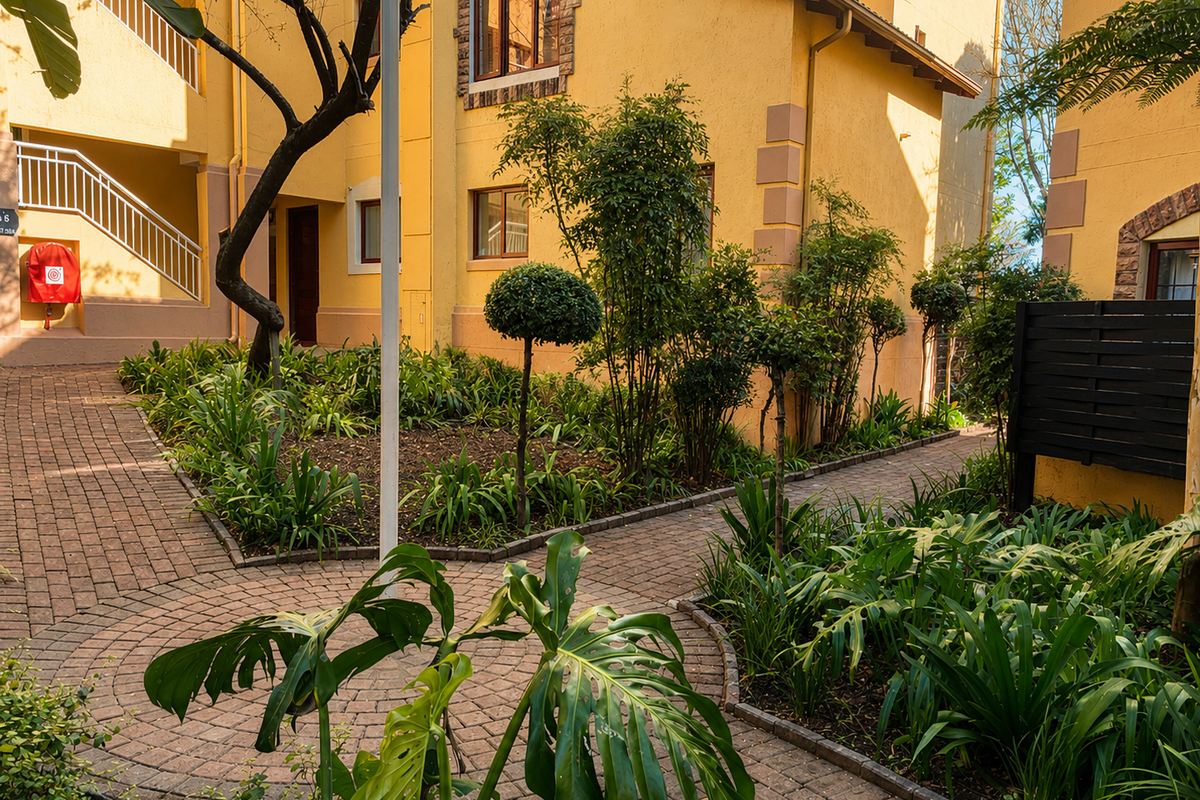 A well-maintained apartment complex courtyard featuring paved walkways, landscaped garden beds, and exterior building access with staircases.