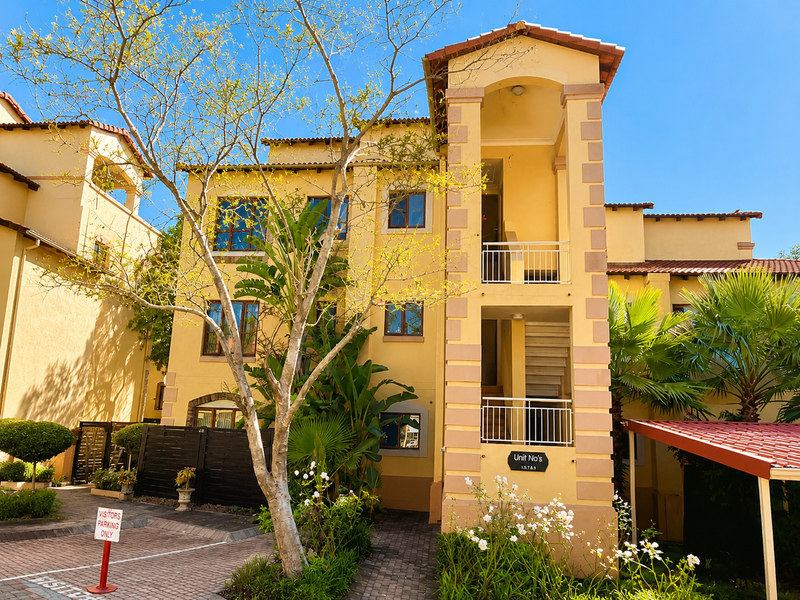 Exterior view of a multi-story apartment complex featuring yellow stucco walls, terracotta roof tiles, and a paved entrance area with covered parking.