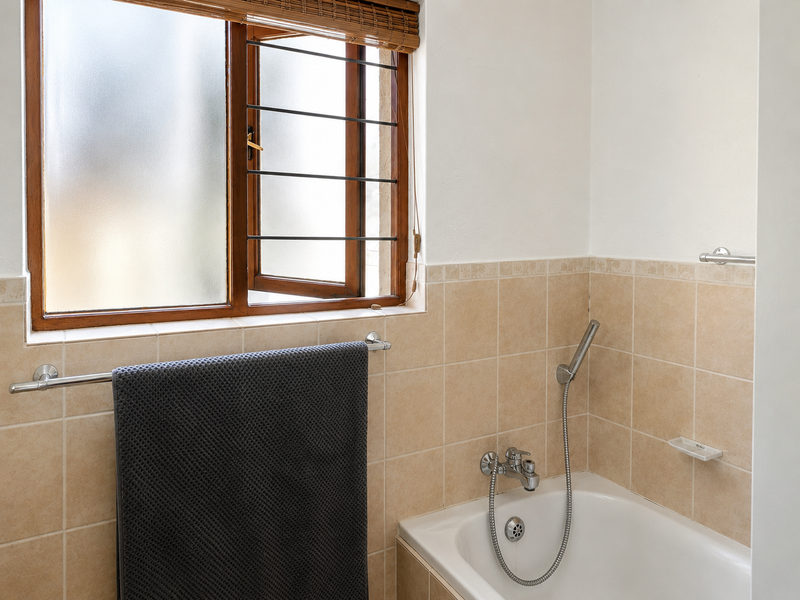 A functional bathroom featuring a bathtub with a handheld shower mixer, tiled wall surrounds, and a timber-framed window with security bars.