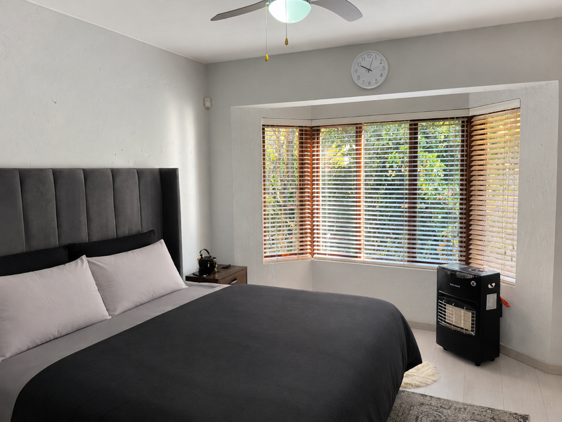 A well-lit bedroom featuring a large bay window with venetian blinds, a ceiling fan, and neutral wall finishes.