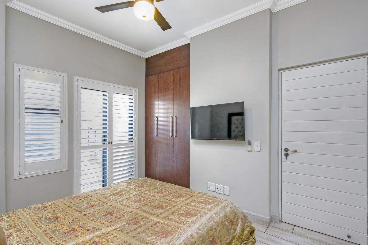 A well-maintained bedroom featuring built-in wooden wardrobes, plantation shutters, a ceiling fan, and neutral tiled flooring.