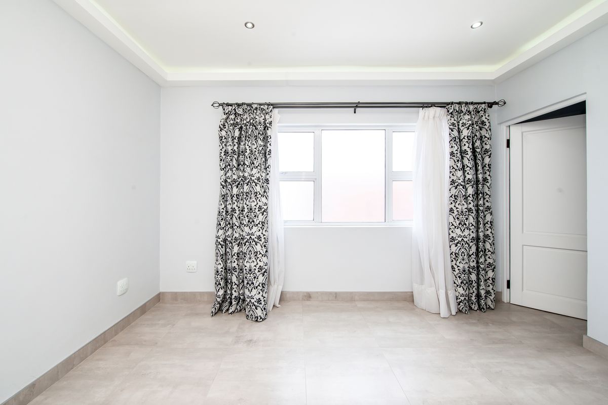A bright, empty bedroom featuring neutral tiled flooring, recessed ceiling lighting, and a large window with curtain fixtures.
