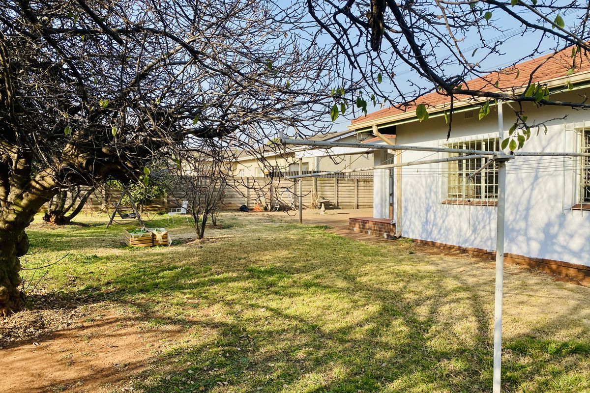 A view of the rear garden featuring a lawn, mature trees, a rotary clothesline, and the rear facade of the house with security bars.