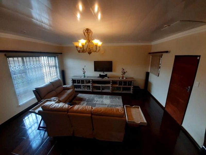 A spacious living room featuring dark wood flooring, a decorative chandelier, and large windows with fitted blinds.