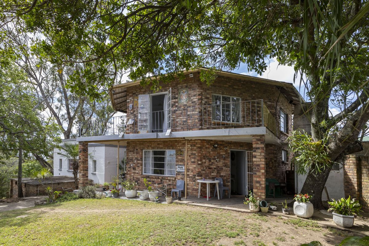 Two-story exposed brick house featuring an upper-level balcony, ground-floor patio, and mature garden surroundings.