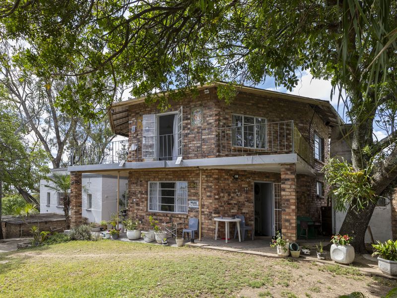 Two-story exposed brick house featuring an upper-level balcony, ground-floor patio, and mature garden surroundings.