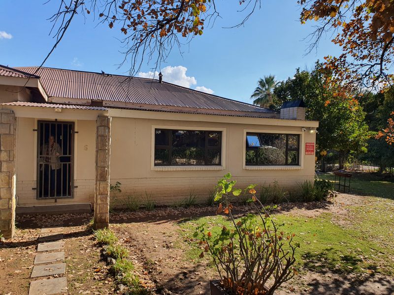 Front elevation of a single-story house featuring a corrugated metal roof, stone-clad entrance pillar, and security gate, set within a garden area.