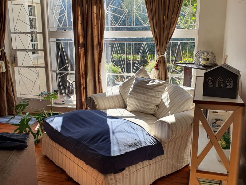 A sunlit living room featuring polished hardwood floors, large windows with security bars, and a wall-mounted split-system air conditioning unit.