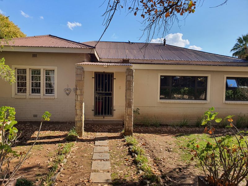 Front elevation of a single-story house featuring a corrugated metal roof, stone-clad entrance pillars, and a paved walkway leading to the main security gate.