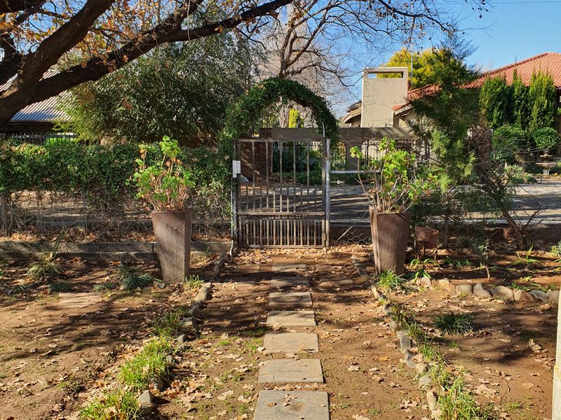 A garden pathway featuring stone pavers leading to a metal gate and archway, surrounded by mature trees and established landscaping.