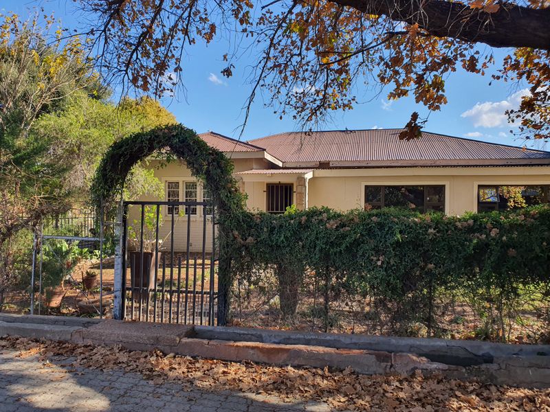 Front view of a single-story house featuring a metal gate entrance, corrugated roof, and security bars on windows, set behind a mature garden.