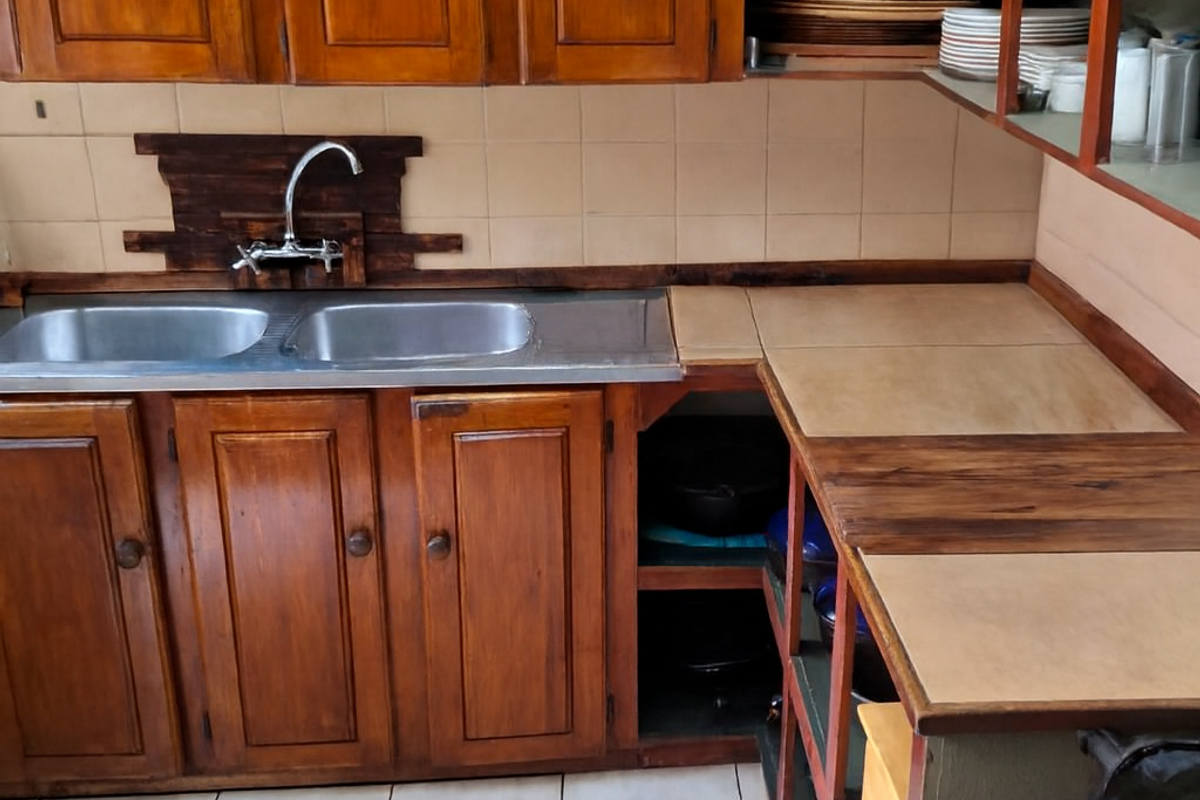 A dated kitchen featuring wooden cabinetry, a double bowl stainless steel sink, tiled splashback, and tiled flooring with open shelving storage.
