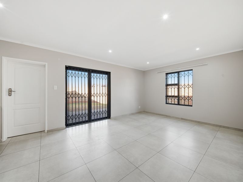 A bright, empty living room featuring neutral tiled flooring, recessed downlighting, and secure access to the exterior via a sliding door with security gate.