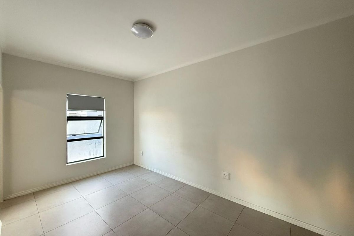 A clean, empty bedroom featuring neutral tiled flooring, a top-hung window with a roller blind, and a simple ceiling light fixture.