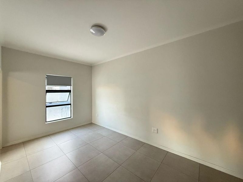 A clean, empty bedroom featuring neutral tiled flooring, a top-hung window with a roller blind, and a simple ceiling light fixture.