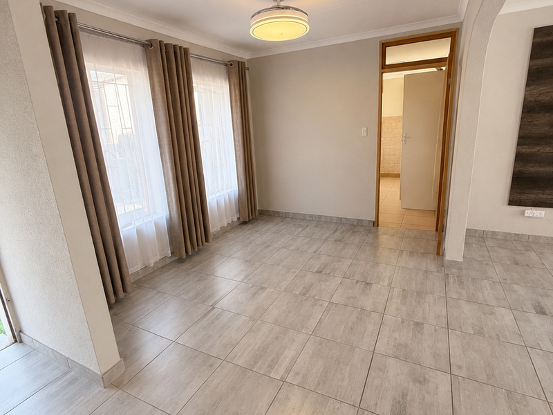 A bright, empty living area featuring neutral tiled flooring, a modern ceiling light, and large windows with security bars, leading into an adjacent room.