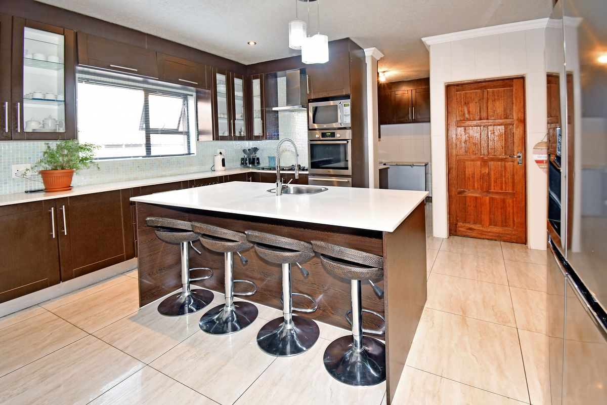 Modern kitchen featuring a central island with a sink, dark wood cabinetry, built-in appliances, mosaic backsplash, and light-toned tiled flooring.
