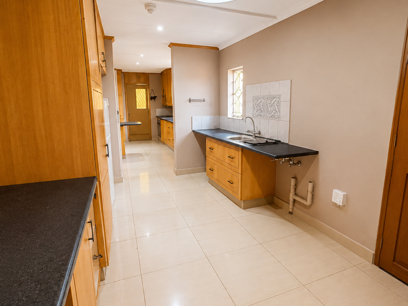 A functional scullery or secondary kitchen area featuring wooden cabinetry, dark countertops, a stainless steel sink, and tiled flooring with a skylight.