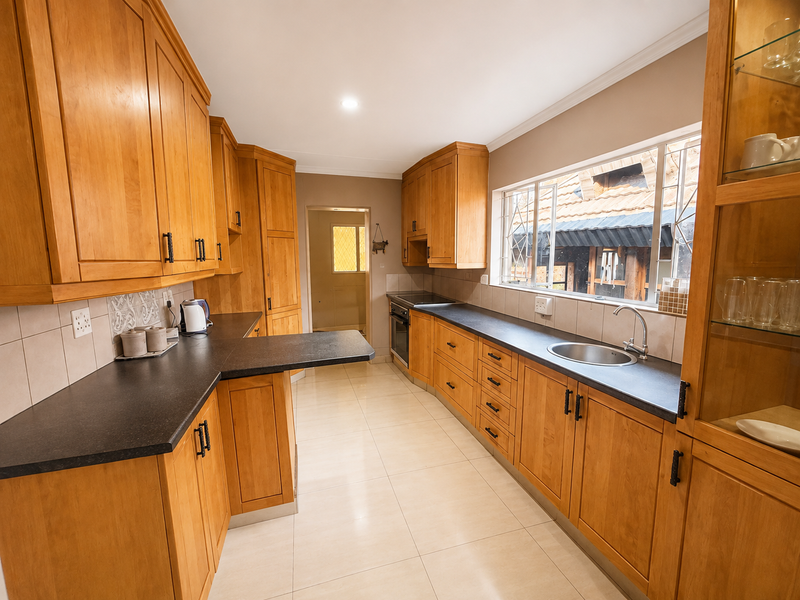 A functional kitchen featuring warm-toned wooden cabinetry, dark laminate countertops, a stainless steel sink, and tiled flooring with a view to an adjacent utility area.