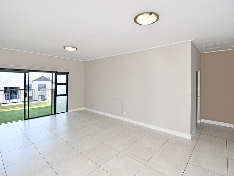 A bright, empty living room featuring neutral ceramic floor tiles, flush mount lighting, and a sliding glass door leading to an outdoor balcony area.