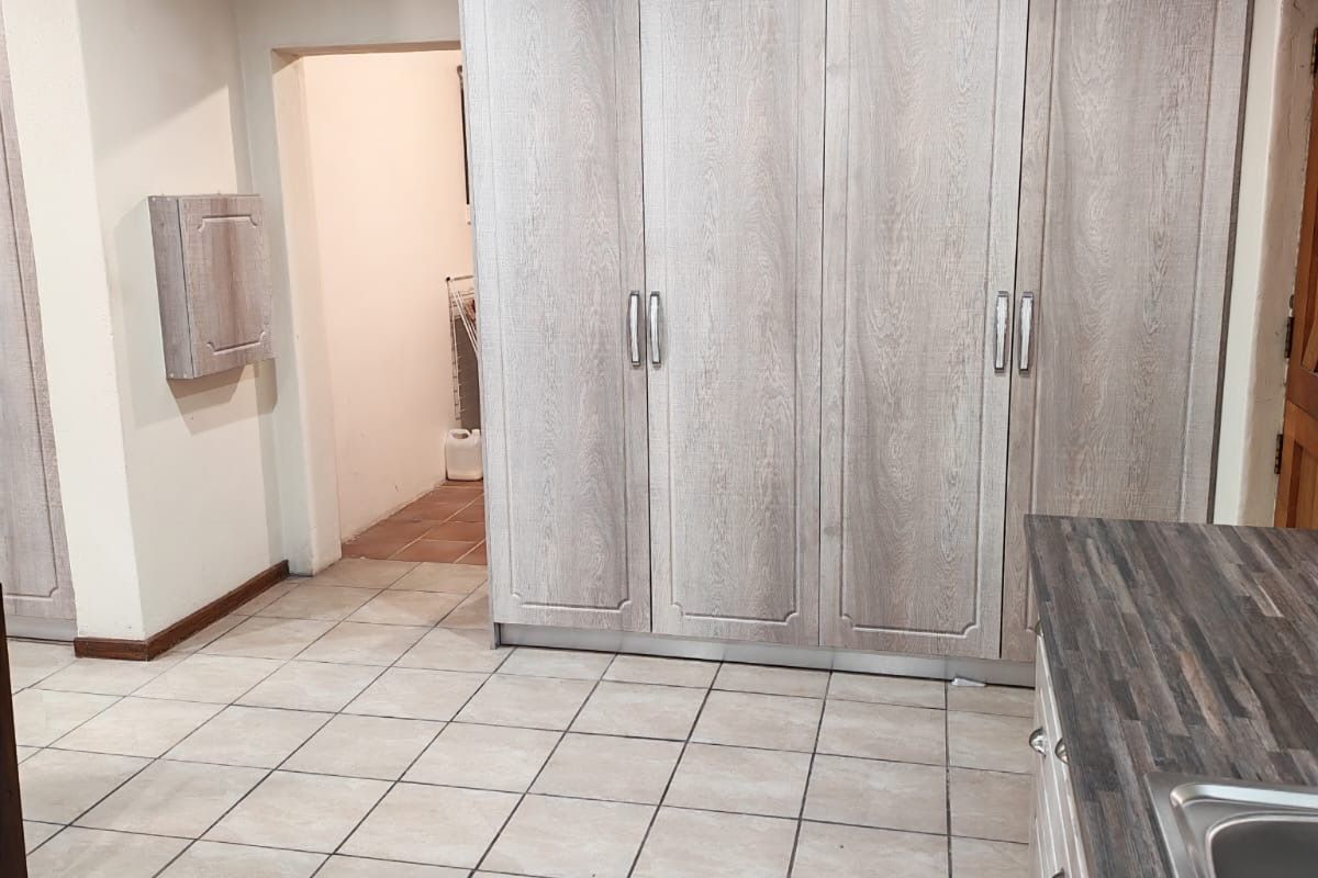 A functional kitchen area featuring light wood-grain cabinetry, dark laminate countertops, a stainless steel sink, and tiled flooring with a doorway leading to a utility space.