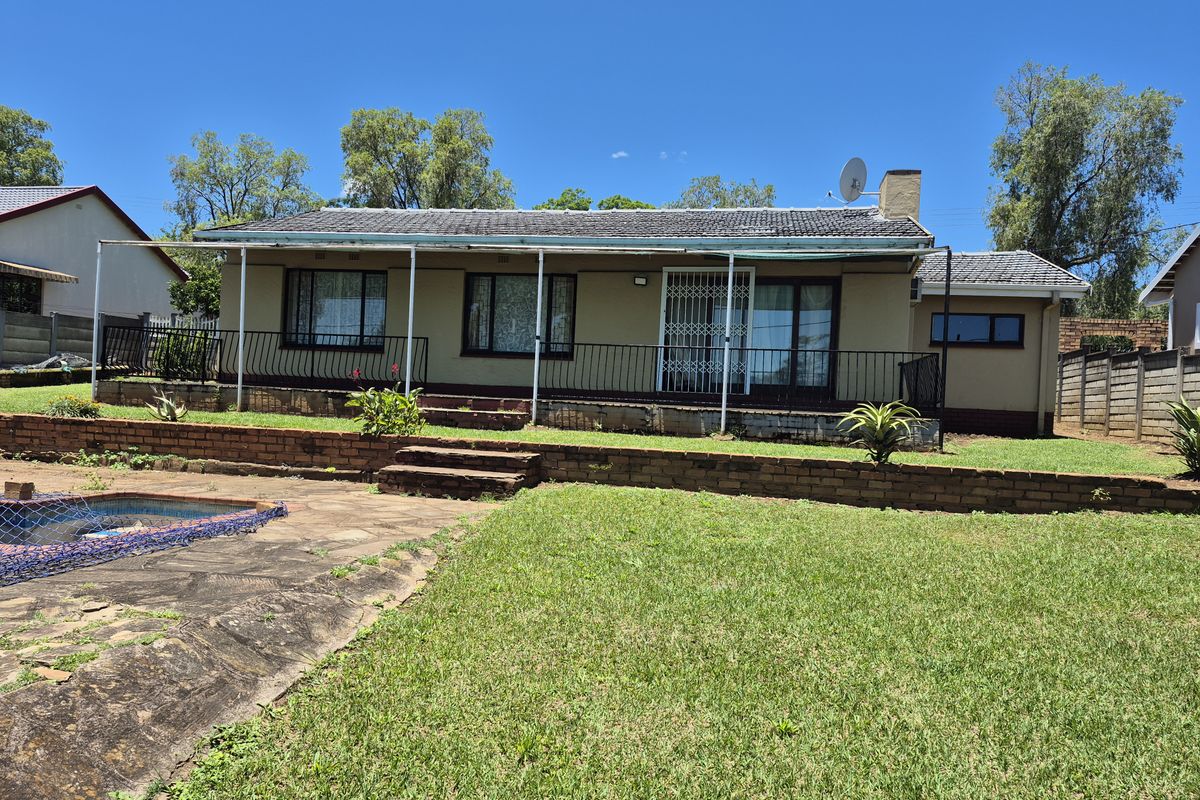 Rear view of a single-story house featuring a covered patio, brick retaining wall, and a swimming pool set within a grassed garden.