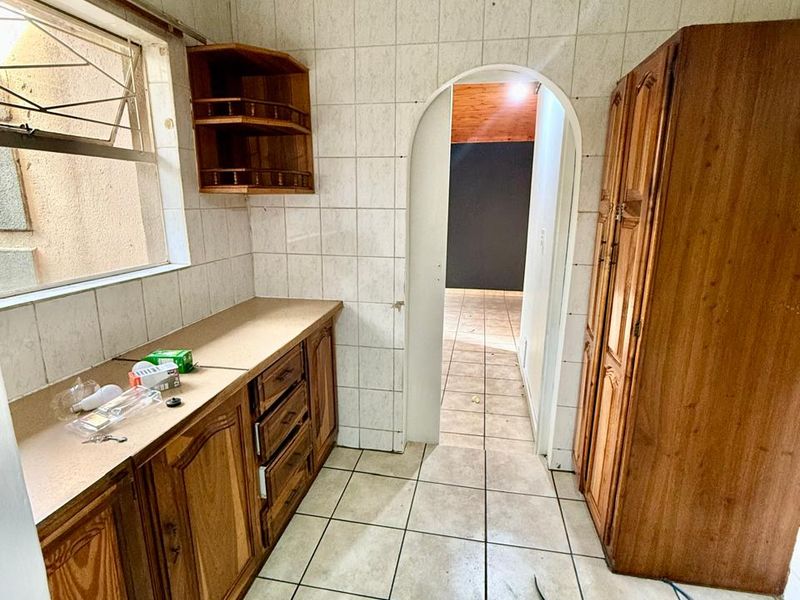 A dated kitchen featuring wooden cabinetry, tiled walls and flooring, and an arched doorway leading to an adjacent living space.