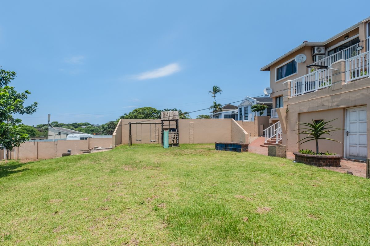 Rear view of the townhouse featuring a spacious lawn, boundary walls, paved patio area, and an elevated balcony with external access.